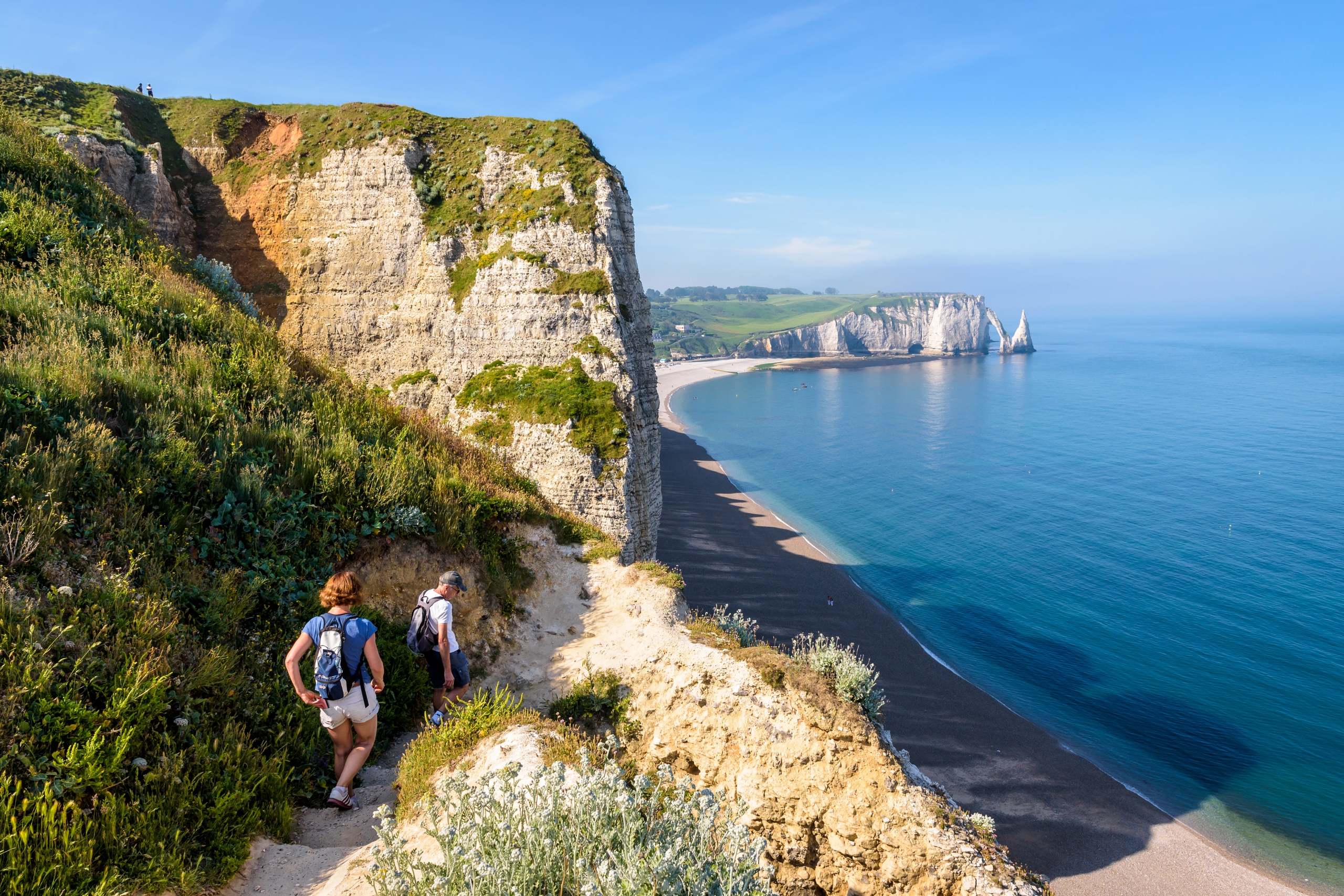 A couple in Normandy, France, walking down the rock stairs to the beach at the foot of the Amont cliff. In the distance the Aval cliff, the arch and the Needle.